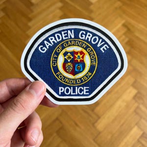 May include: A blue and white Garden Grove Police patch. The patch features the words "Garden Grove" and "Police" in white lettering. The center emblem includes a shield with a star and other symbols. The patch is edged with a white border.