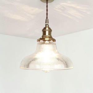 May include: A vintage-style pendant light fixture with a clear, ribbed glass shade and brass-colored hardware. The light hangs from a brown cord and is mounted on a brass-colored ceiling plate. The glass shade is shaped like a shallow bowl.
