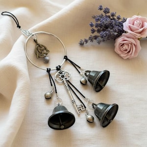 May include: A silver-toned wind chime featuring a large ring with decorative charms and three small bells. The charms include a Celtic knot, a key, and small bell-shaped accents. The bells are dark gray and suspended by black cords.