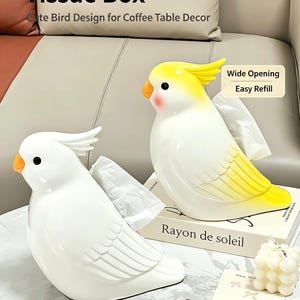 May include: Two parrot-shaped ceramic tissue boxes. One is solid white with an orange beak, and the other is white with a yellow head, crest, and tail, featuring an orange beak and pink cheeks. Both have a wide opening for tissue dispensing.