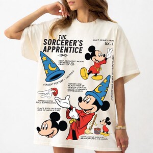 Può includere: Una t-shirt color crema con una stampa grafica de L'Apprendista Stregone del 1940. Il design include illustrazioni di Topolino con una tunica rossa e un cappello da mago blu, insieme a note tecniche e testo che descrive le caratteristiche.