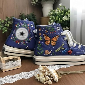 Könnte beinhalten: Ein Paar blaue High-Top-Sneaker aus Segeltuch mit detaillierten Stickereien eines Monarchfalters, einer Gottesanbeterin, einer Schnecke, eines Marienkäfers sowie kleiner Blumen und Beeren. Die Schuhe haben weiße Schnürsenkel und eine weiße Sohle.