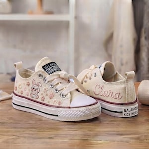 May include: A pair of cream-coloured canvas trainers featuring a white rubber toe cap and sole with a black stripe. The left shoe displays an embroidered rabbit and small flowers. The right shoe features the name Clara in pink script embroidery.