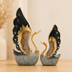 May include: Two decorative swan figurines. The swans have gold-coloured necks and beaks, with intricately designed wings in black and gold. The bases are patterned with a floral design in shades of grey and teal. The swans are displayed on a wooden surface.