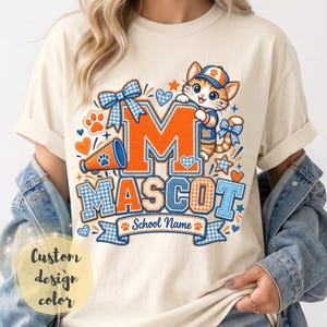 May include: A cream-coloured t-shirt featuring a graphic of a cartoon cat wearing a baseball cap and jersey. The design includes a large orange letter M, the word MASCOT, a megaphone, blue gingham bows, and a banner with the text School Name.