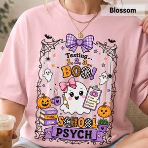 May include: A light pink t-shirt featuring a graphic with a ghost wearing a pink bow, holding a pencil and a clipboard. The text reads Testing... 1, 2, 3... BOO! SCHOOL PSYCH. Includes pumpkins, books labeled Feelings, Think, Grow, and spiderwebs.