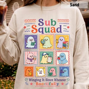 May include: A sand-colored crewneck sweatshirt featuring a colorful graphic print with the text Sub Squad and Winging It Since Whenever Boo-tifully. The design includes nine square panels, each containing a cartoon ghost with various school-themed items.