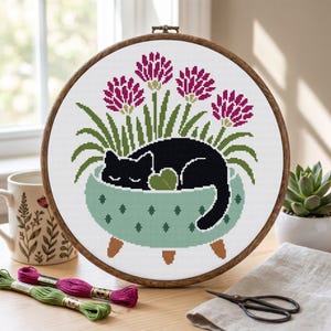Könnte beinhalten: Ein runder Holzstickrahmen mit einem Kreuzstichmuster, das eine schwarze Katze zeigt, die in einem hellgrünen, dreibeinigen Pflanzgefäß schläft. Das Design zeigt vier magentafarbene Blumen mit grünen Stielen und Blättern auf weißem Stoff.