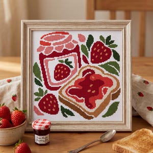 May include: A framed cross-stitch embroidery featuring a red strawberry jam jar, a slice of toast with red jam, and fresh strawberries. The design uses red, pink, green, and brown thread on white fabric, enclosed in a light wood-toned frame.