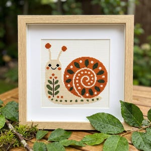May include: A framed cross-stitch artwork featuring a whimsical snail design. The snail has a cream-colored body with orange and green shell details. The artwork is framed in a light wood frame and set against a wooden surface.
