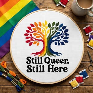 Könnte beinhalten: Ein runder hölzerner Stickrahmen mit weißem Stoff, der ein im Kreuzstich gesticktes Regenbogenbaum-Motiv zeigt. Unter dem Baum steht der Text Still Queer, Still Here in schwarzen Blockbuchstaben. Das Design zeigt ein Farbspektrum.
