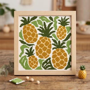 May include: A square cross-stitch embroidery featuring a pattern of four pineapples in shades of yellow and gold with dark green leaves, set within a light-colored wooden frame. The design is displayed on a white fabric background.