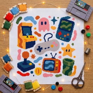 Peut inclure: Un tissu de point de croix blanc présentant des icônes de jeux vidéo pixelisées colorées, dont une borne d'arcade, une console portable, une manette, un fantôme, une étoile, une télévision et un tuyau, entouré de bobines de fil et de cartouches.
