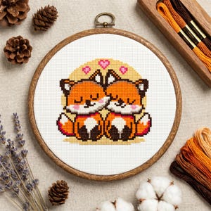 May include: A circular wooden embroidery hoop containing a cross-stitch pattern of two orange foxes with white chests and black ears, sitting side-by-side with closed eyes. Three small pink hearts are stitched above the foxes on a yellow background.