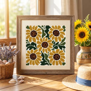 May include: A square framed cross-stitch artwork featuring a pattern of four yellow sunflowers with dark brown centers and dark green leaves, set against a white background, enclosed in a light brown wood-grain textured frame with a cream mat.