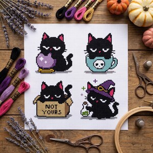 May include: A cross-stitch pattern on white fabric featuring four black cats. One cat holds a purple crystal ball, one sits in a skull-patterned mug, one sits in a box labelled NOT YOURS, and one wears a purple witch hat while holding a green potion.