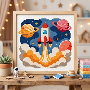 May include: A framed cross-stitch embroidery featuring a white and red rocket ship launching into a dark blue space background with yellow stars, a yellow ringed planet, a pink ringed planet, and a red planet, all rising from orange smoke.
