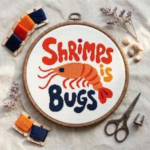 May include: A circular wooden embroidery hoop containing white Aida fabric with a cross-stitch design of an orange shrimp and the text Shrimps is Bugs in red and dark blue. Includes embroidery floss bobbins, metal scissors, and a thimble.