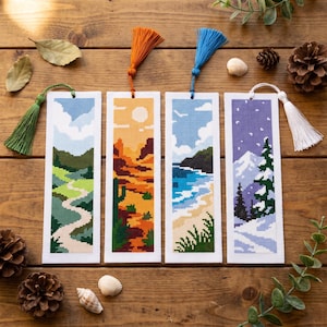 May include: Four rectangular cross-stitch bookmarks featuring seasonal landscapes. The first shows a green path, the second a desert with a cactus, the third a coastal beach, and the fourth a snowy mountain scene with pine trees. Each has a tassel.