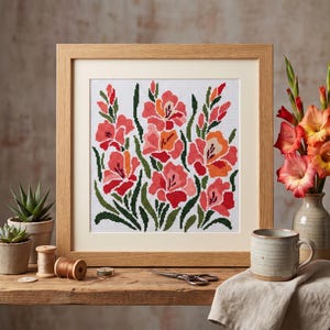 May include: A framed cross-stitch embroidery featuring a pattern of pink, red, and orange gladiolus flowers with green leaves on a white grid background, encased in a light-colored wooden frame with a cream-colored mat board border.