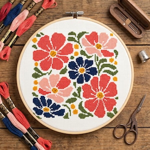 Peut inclure: Un tambour à broder circulaire en bois contenant un motif au point de croix de fleurs rouges, roses et bleu foncé avec des feuilles vertes sur tissu blanc. Autour du tambour se trouvent des échevettes de fil, un étui à aiguilles et des ciseaux.