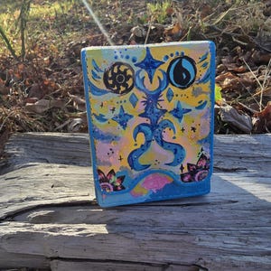 May include: A rectangular deck box featuring a hand-painted design with a yellow, pink, and blue background. The front displays dark blue abstract symbols, including a sun and a water drop icon, surrounded by star shapes and floral motifs.