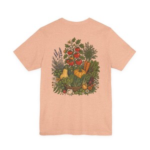 Puede incluir: Camiseta color melocotón con una ilustración detallada de una cosecha de jardín. El diseño incluye tomates rojos maduros, una calabaza amarilla, zanahorias y otras verduras y hierbas, todo dispuesto en una cesta.