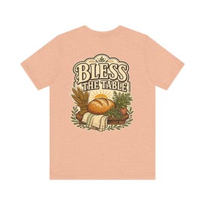 以下が含まれることがあります： ヴィンテージスタイルのグラフィックデザインで「BLESS THE TABLE」の文字が入った、ピーチ色のTシャツ。パン、小麦、ハーブ、朝日が、ストライプの布が敷かれた木製のテーブルに配置されています。