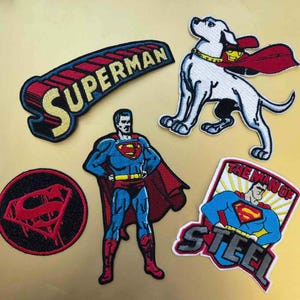 Peut inclure: Écussons brodés représentant Superman et son logo. Comprend un écusson avec le mot "SUPERMAN", un écusson du chien de Superman et des écussons avec Superman dans des poses d'action. Les écussons sont rouges, bleus, jaunes et noirs.