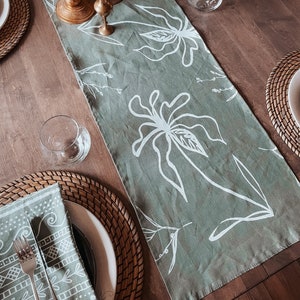 Può includere: Un runner da tavolo verde con un motivo floreale bianco. Il runner è disposto su un tavolo di legno con posate, tra cui tovagliette all'uncinetto, piatti bianchi e bicchieri.