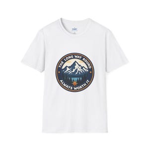 Puede incluir: Camiseta blanca con un gráfico circular que representa una cordillera, un lago y el texto "THE LONG WAY ROUND ALWAYS WORTH IT". El gráfico es en tonos azules, beige y marrón. La camiseta tiene cuello redondo y mangas cortas.