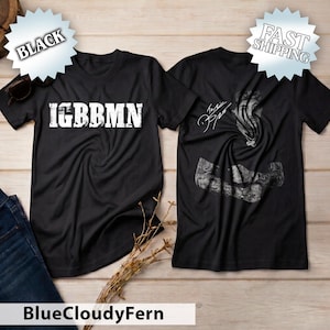 May include: A black short-sleeve t-shirt featuring the white text IGBBMN on the front. The back of the shirt displays a black and white graphic of a person with tattoos and a signature. The shirt is presented in a flat lay view.