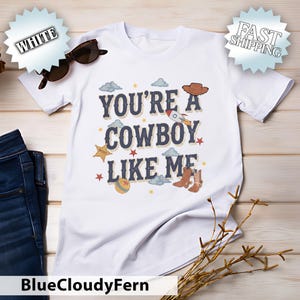 May include: A white short-sleeved t-shirt featuring the text "YOU'RE A COWBOY LIKE ME" in dark blue letters. The design includes illustrations of a brown cowboy hat, a rocket, a sheriff star badge, a ball, and cowboy boots with small clouds and stars.