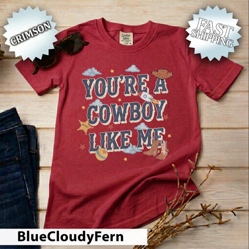 May include: A crimson short-sleeve t-shirt featuring the text "YOU'RE A COWBOY LIKE ME" in dark blue letters. The design includes illustrations of a cowboy hat, a rocket, a sheriff star, a ball, and cowboy boots, accented with light blue clouds.