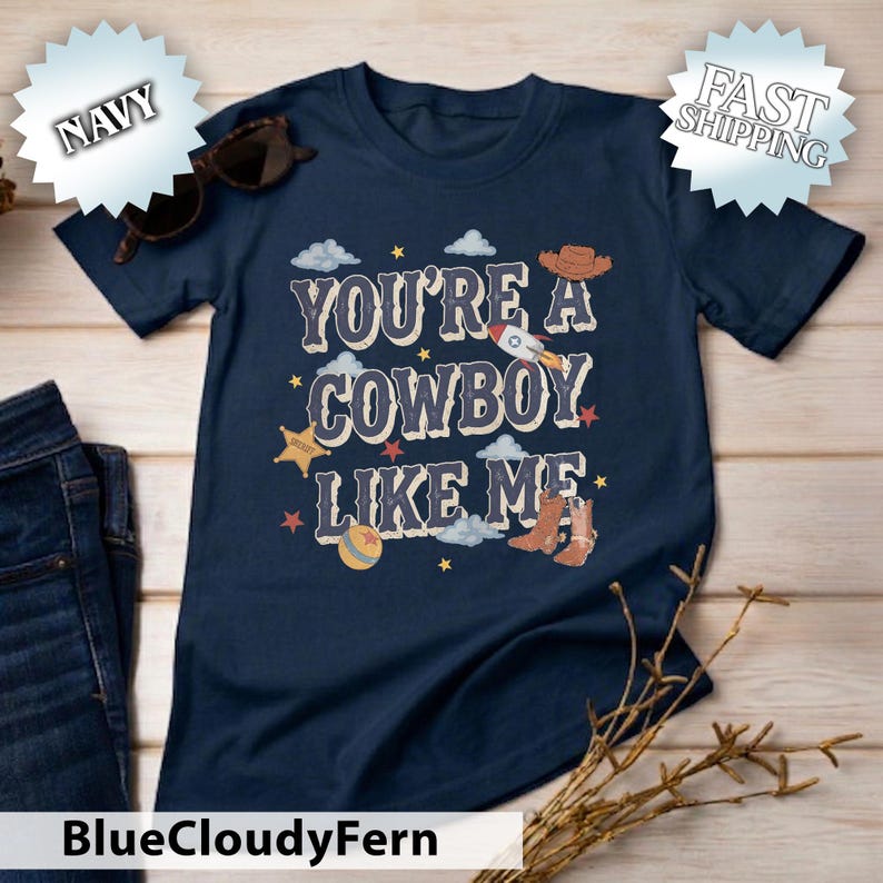 May include: A navy blue short-sleeved t-shirt featuring the text "YOU'RE A COWBOY LIKE ME" in a light-coloured font. The graphic includes illustrations of a rocket, a cowboy hat, a sheriff star badge, a ball, and a pair of brown cowboy boots.