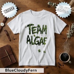 May include: A white short-sleeved t-shirt featuring the text "TEAM ALGAE" printed in a stylized, dripping green font that resembles slimy algae. The graphic is centered on the chest of the shirt, which has a crew neckline and a standard fit.