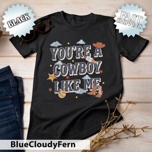 May include: A black short-sleeved t-shirt featuring the text "YOU'RE A COWBOY LIKE ME" in a distressed font. The graphic includes illustrations of a rocket, a cowboy hat, a sheriff star, a ball, and a pair of brown cowboy boots with clouds and stars.