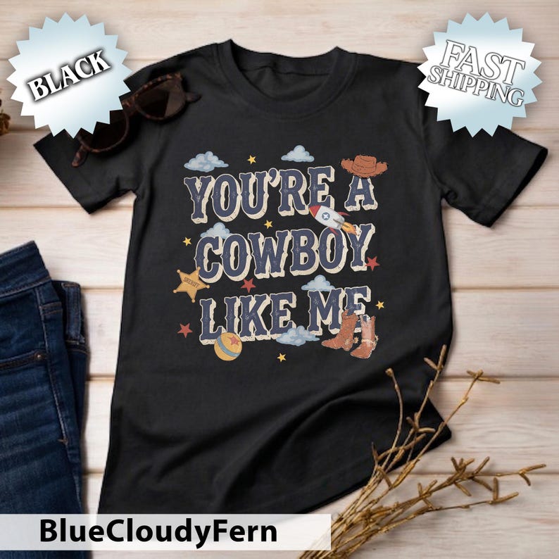 May include: A black short-sleeved t-shirt featuring the text "YOU'RE A COWBOY LIKE ME" in a distressed font. The graphic includes illustrations of a rocket, a cowboy hat, a sheriff star, a ball, and a pair of brown cowboy boots with clouds and stars.
