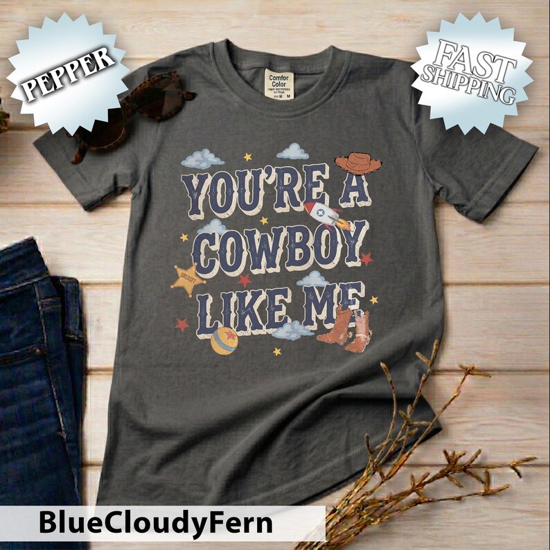 May include: A charcoal grey short-sleeve t-shirt featuring the text "YOU'RE A COWBOY LIKE ME" in a bold font. The graphic includes illustrations of a cowboy hat, a rocket, a sheriff star, a ball, and cowboy boots, surrounded by small stars and clouds.