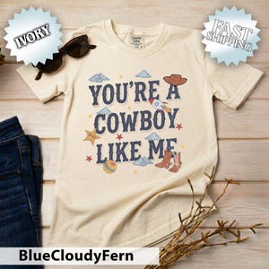 May include: An ivory short-sleeve t-shirt featuring the text "YOU'RE A COWBOY LIKE ME" in dark blue letters. The design includes illustrations of a brown cowboy hat, a rocket ship, a sheriff star, a ball, and brown cowboy boots with clouds.
