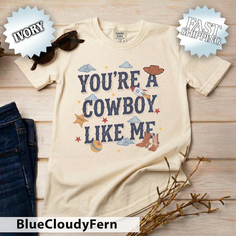 May include: An ivory short-sleeve t-shirt featuring the text "YOU'RE A COWBOY LIKE ME" in dark blue letters. The design includes illustrations of a brown cowboy hat, a rocket ship, a sheriff star, a ball, and brown cowboy boots with clouds.