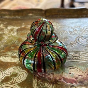 May include: A small, round glass perfume bottle featuring a ribbed design with alternating vertical stripes of red, green and gold. The bottle includes a matching spherical glass stopper that sits securely in the centre of the rounded base.
