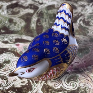May include: A porcelain bird figurine featuring a deep blue body with gold leaf-shaped patterns, a white head with a red eye, and gold-trimmed wings with white and blue scale details. The tail feathers are white with blue and gold accents.