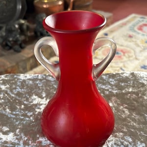 May include: A vibrant red glass vase featuring a bulbous base, a flared rim, and two clear, curved glass handles on opposite sides. The surface of the red glass has a smooth, matte finish, while the handles are transparent and smooth.