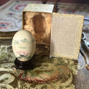 May include: An off-white egg-shaped ornament featuring a hand-painted landscape with green mountains and pink flowering trees. The egg rests on a dark brown circular wooden stand and is accompanied by an open, gold-patterned storage box.