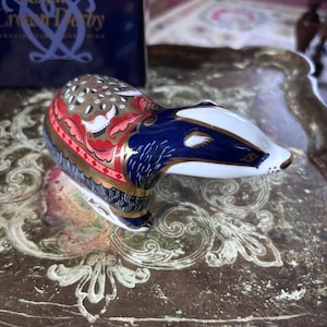 Peut inclure: Une figurine de blaireau en porcelaine Royal Crown Derby avec une tête blanche aux marques noires, un corps bleu profond et des motifs floraux et géométriques complexes dorés, rouges et noirs sur le dos, placée devant sa boîte bleue d'origine.