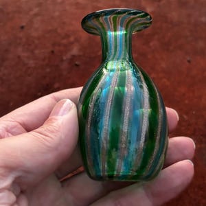 May include: A small glass vase with a rounded body and a narrow neck, featuring vertical stripes in shades of emerald green, teal, and shimmering metallic copper. The glass has a smooth, polished finish with a translucent quality throughout the piece.