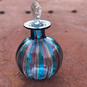 Könnte beinhalten: Eine kleine, runde Parfümflasche aus Glas mit vertikalen Streifen aus durchscheinendem blaugrünem und klarem Glas, akzentuiert durch dünne Bänder aus schimmerndem Goldglitter. Die Flasche hat einen schmalen Hals und einen klaren, blattförmigen Glasstopfen.