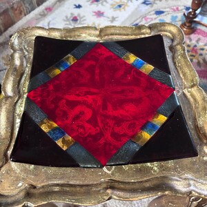 May include: A square glass plate featuring a central red diamond with an intricate scrollwork pattern. The plate is bordered by black glass sections and accented with small, rectangular segments of blue and gold metallic-toned glass.