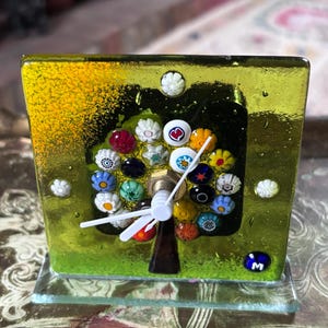 Könnte beinhalten: Eine quadratische Schreibtischuhr aus Glas mit limettengrünem Hintergrund und einem Baummotiv aus bunten Millefiori-Glasperlen. Die Uhr hat weiße Zeiger, einen dunkelbraunen Stamm und einen kleinen blauen runden Akzent mit dem Buchstaben M.